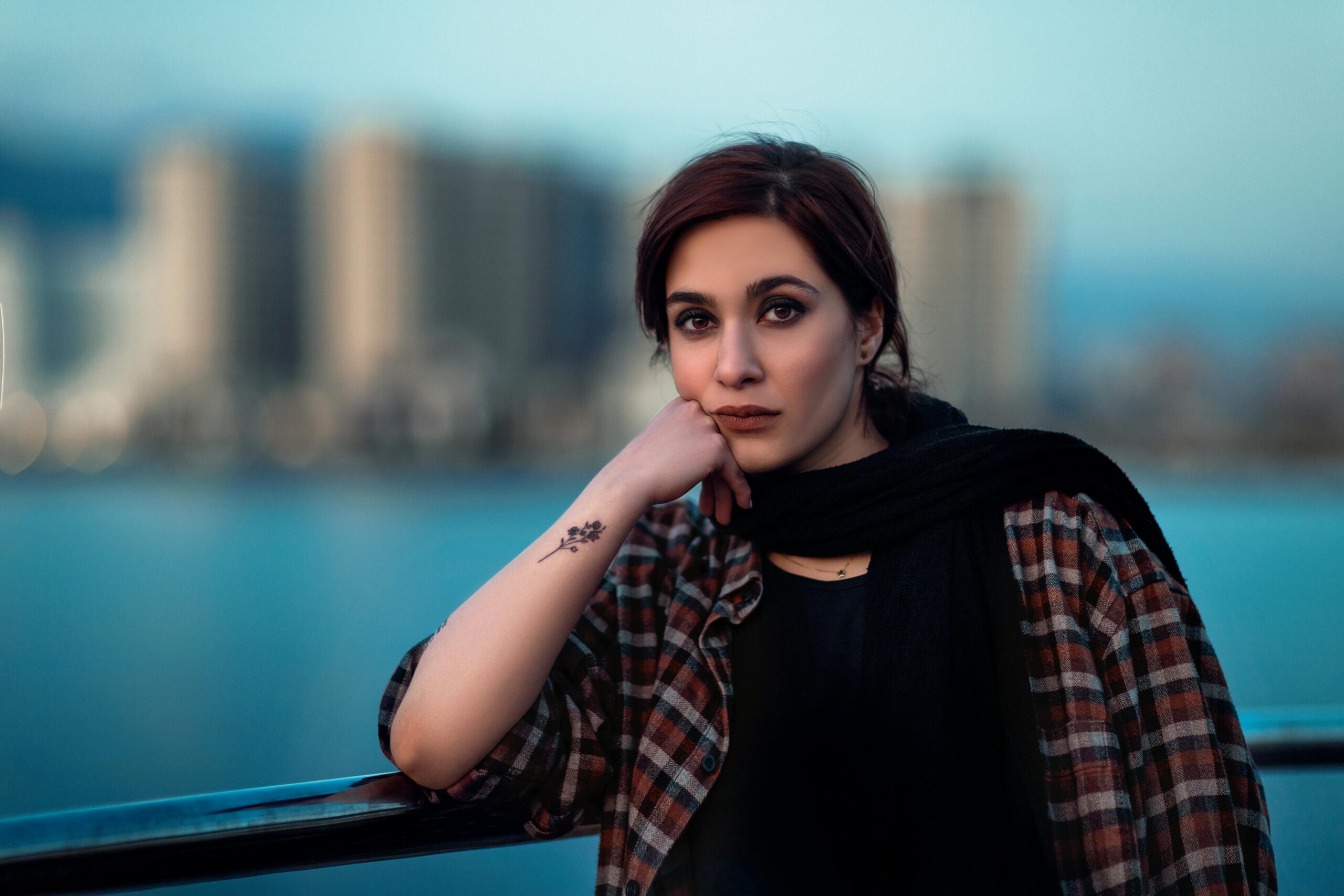 Portrait of a thoughtful woman leaning on a railing by the waterfront with a city backdrop.